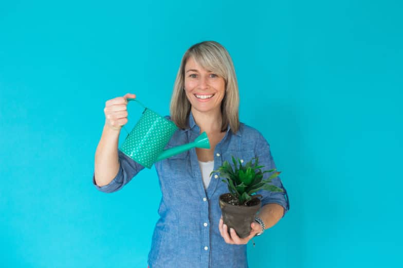 a woman standing in front of a bright blue background uses a spotted watering can to water a small house plant