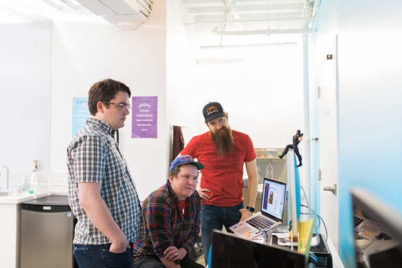 Three men look at a screen in an office setting. Two of them are standing behind the third, who sits in a chair