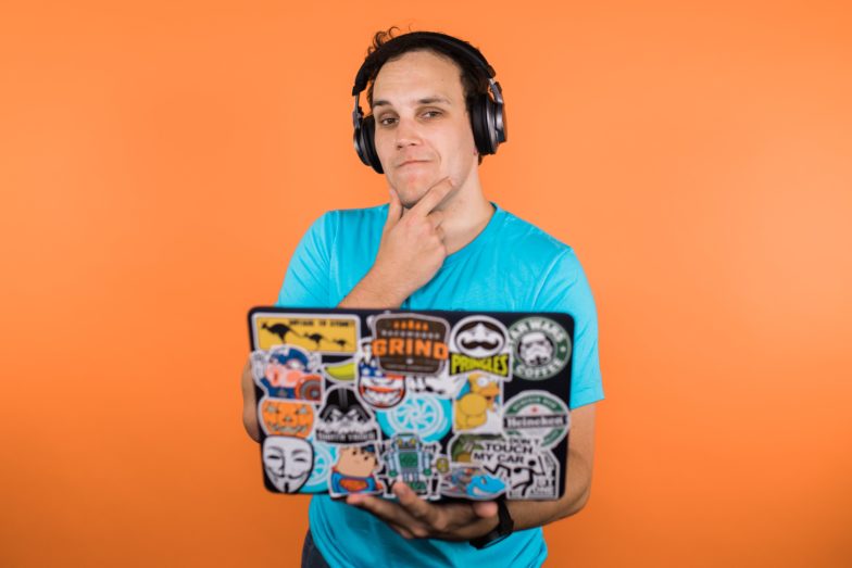 a man holds a computer in front of an orange background. he holds his chin pensively
