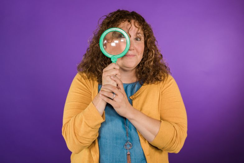 a woman stands in front of a purple background looking through a magnifying glass