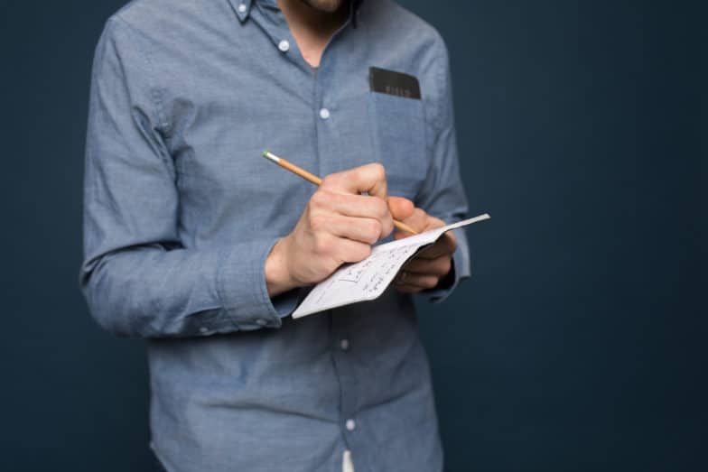 a man standing in front of a navy blue background writes in a notebook with a pencil