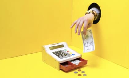 A small cash register in a yellow room is open, displaying the money inside. A hand reached through a hole in the right wall, taking $5 from the till
