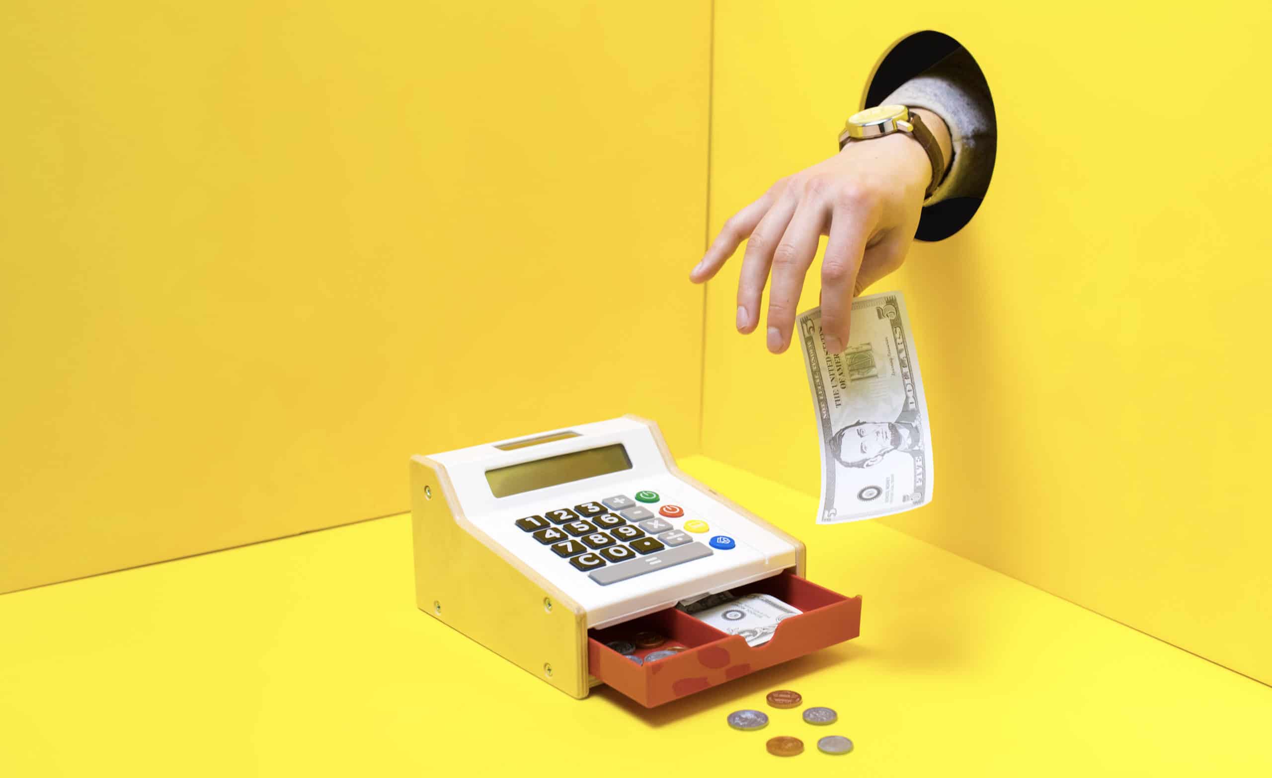 A small cash register in a yellow room is open, displaying the money inside. A hand reached through a hole in the right wall, taking $5 from the till