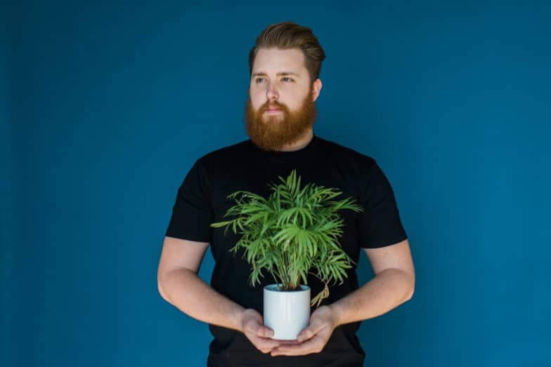 a bearded man in a black shirt holds a house plant in front of a dark blue background