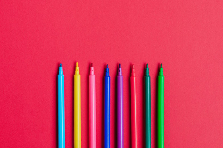 8 different colored markers laid next to one another with the caps off with a red background