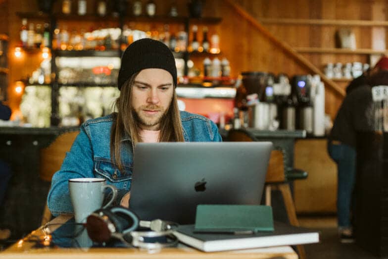 a man wearing a jean jacket and a beanie works on a computer at a coffee shop