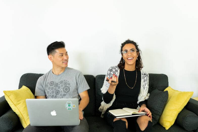 Two people sit on a couch looking at a computer and a notebook 