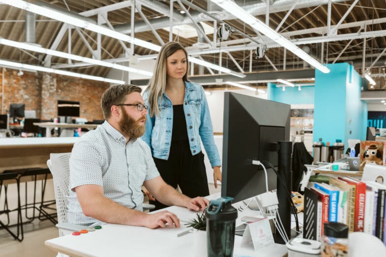 a man sits at a desk and a woman in a jean jacket stands slightly behind him. They both look at a computer screen as they collaborate on a project