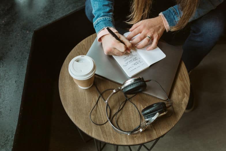 woman writes in small notebook on a table that also holds a laptop, coffee cup, and headphones