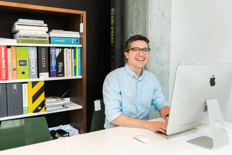 Man sits at desk in front of a monitor by a book case 