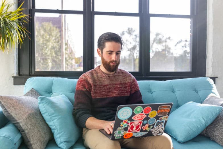 man in sweater sits on blue couch while using a laptop