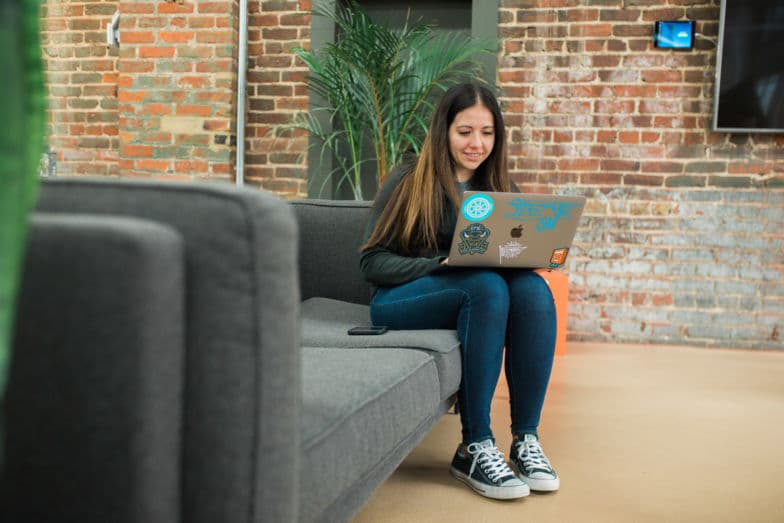 a woman sitting on a gray couch works on a laptop