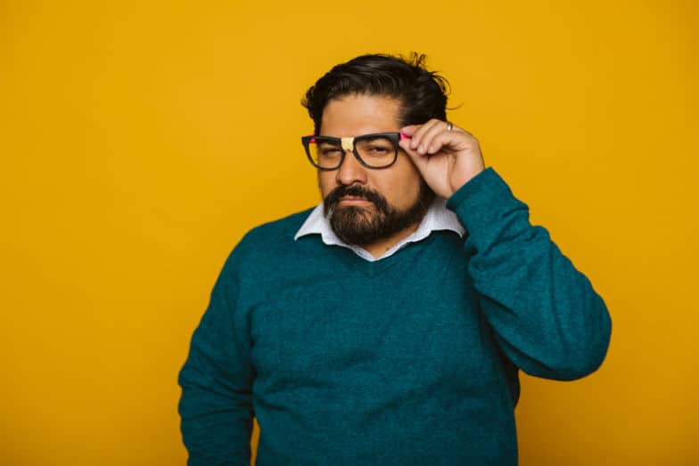 a man peers through black framed glasses in front of a yellow background