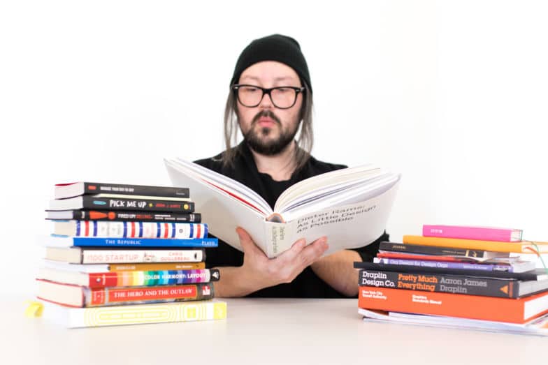 a man in a beanie reads a book on design. two stacks of books sit on either side of his desk