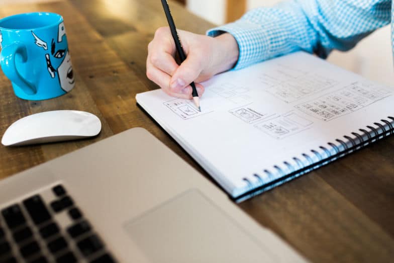 A person sketches a design in a notebook next to a Captain America coffee mug