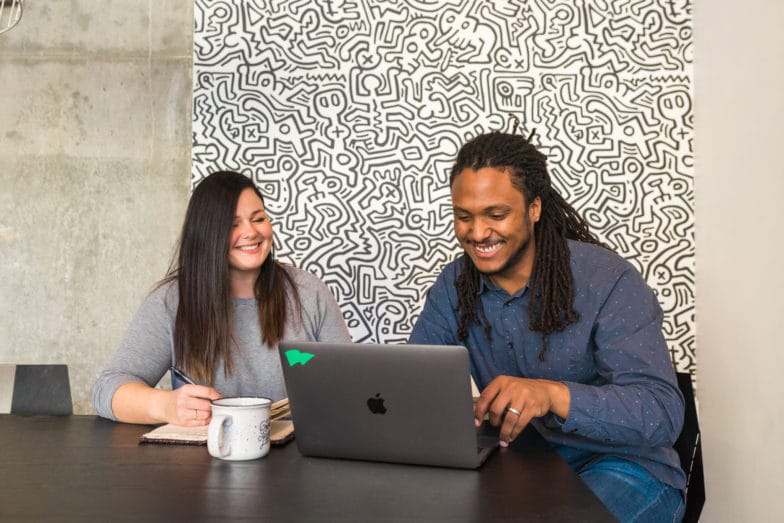 a man and a woman smile at a computer screen while taking a virtual meeting in front of an elaborately detailed black and white wall
