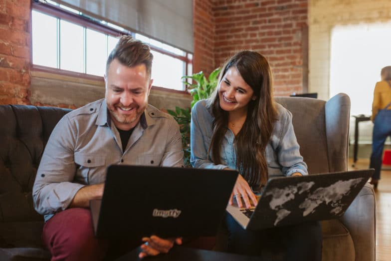 a man and a woman sit on a couch, each holding a laptop. The woman looks over the man's shoulder to see what he's working on as they both smile
