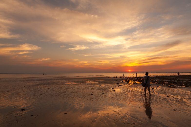 Stock image of a photographer at sunset setting up a tripod on Coca Mocha brown sand to capture images of a cloudy, Perfectly Pale beige sunset