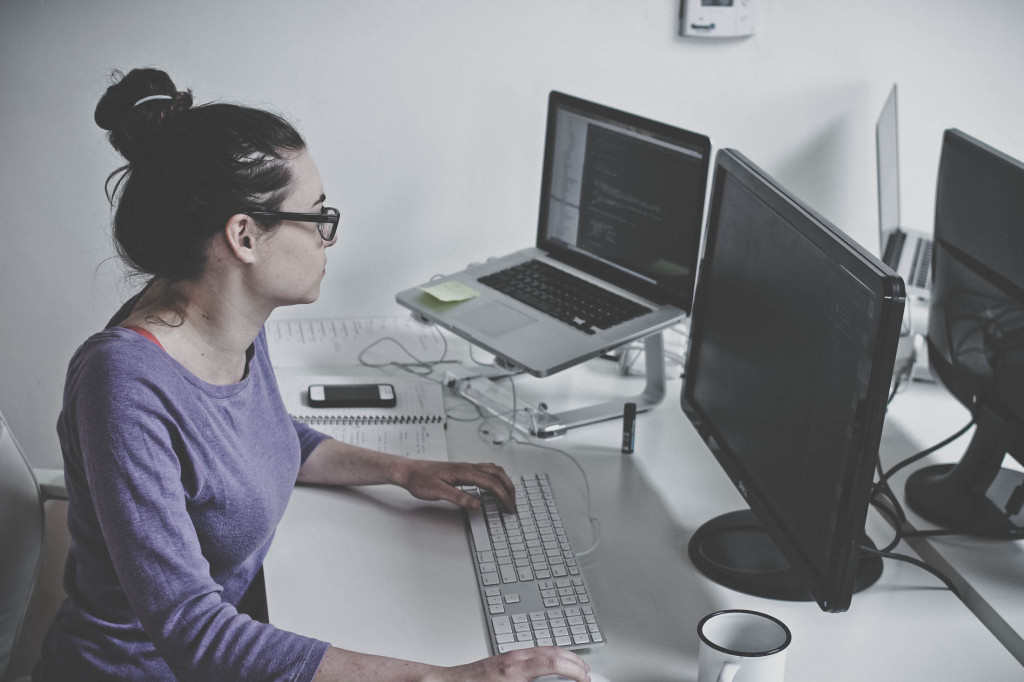 Woman works at a desk in front of two monitors alongside a coffee cup, phone, and notebook.