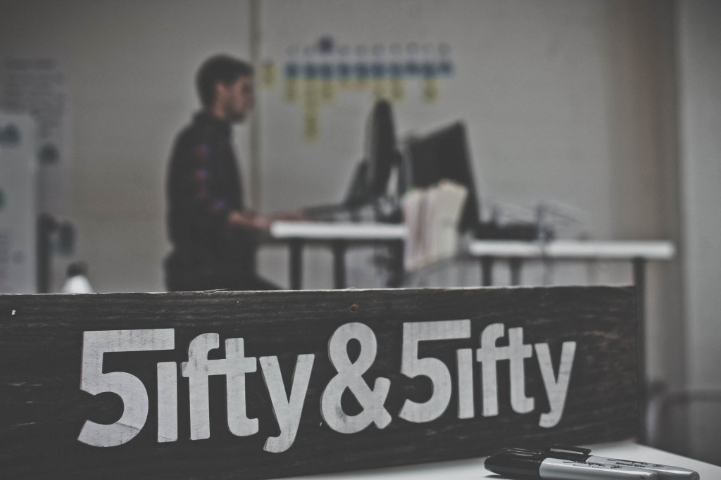 wooden board emblazoned reading Fifty & Fifty written as "5ifty & 5ifty" in white letters. man works on computer in the background