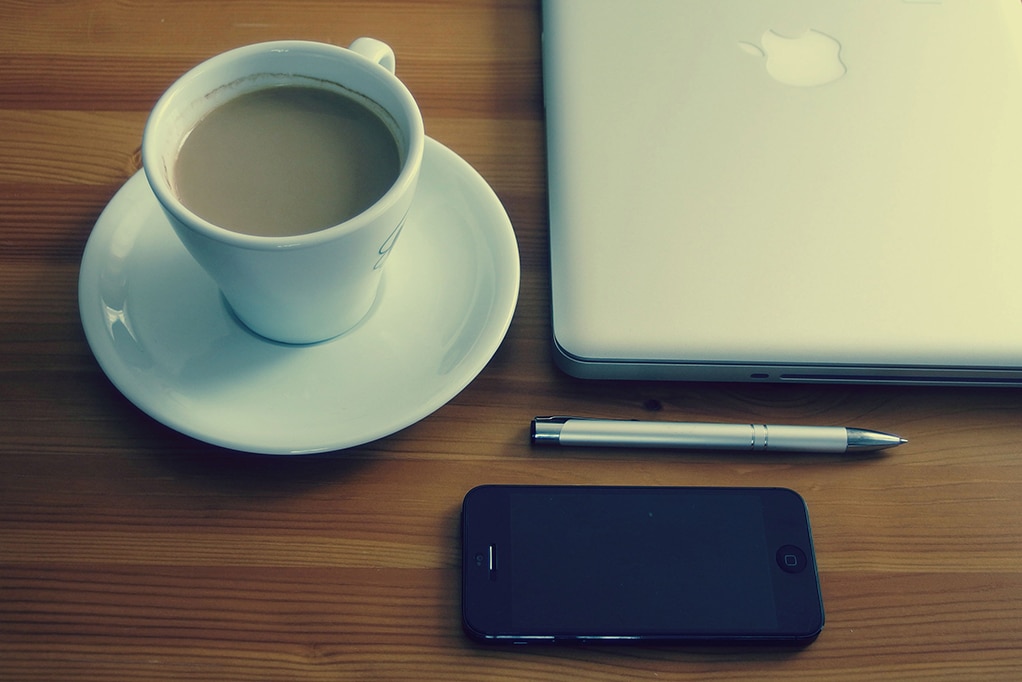 desk with a cup of coffee, a phone, a mac, and a pen