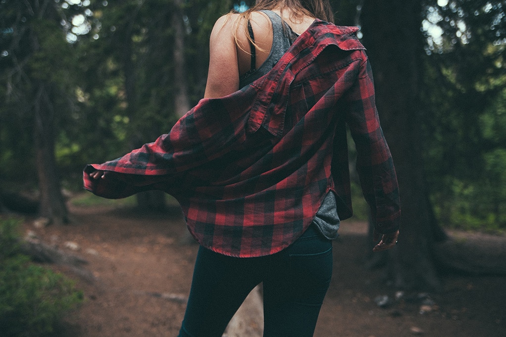 woman on a hike starts to take off a red flannel shirt, leaving the grey tank top beneath it