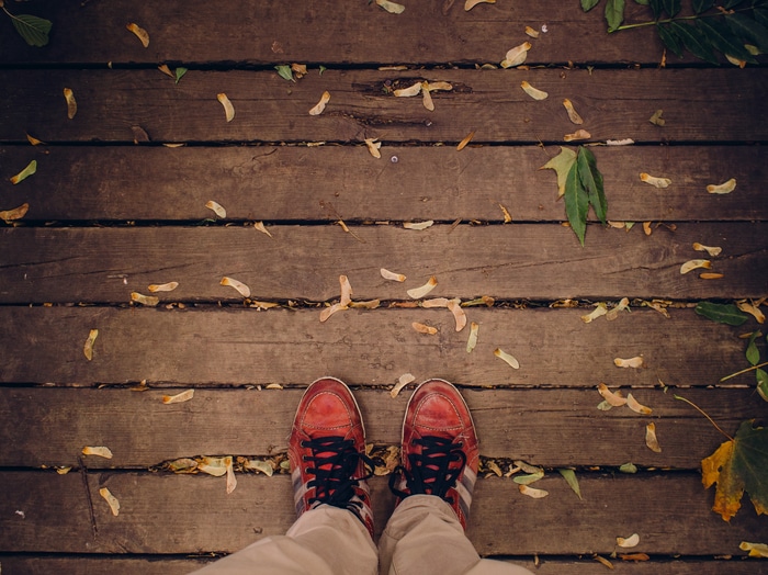 a shot of the ground. Wooden planks run lengthwise across the image, and a person's red shoes and khaki pants are visible