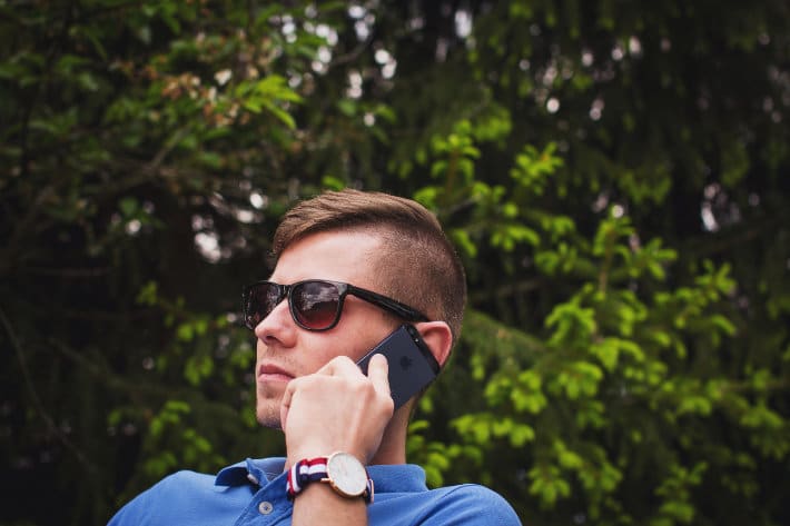 Man in sunglasses and a blue shirt holds a cellphone to his ear in front of a lush, leafy backdrop