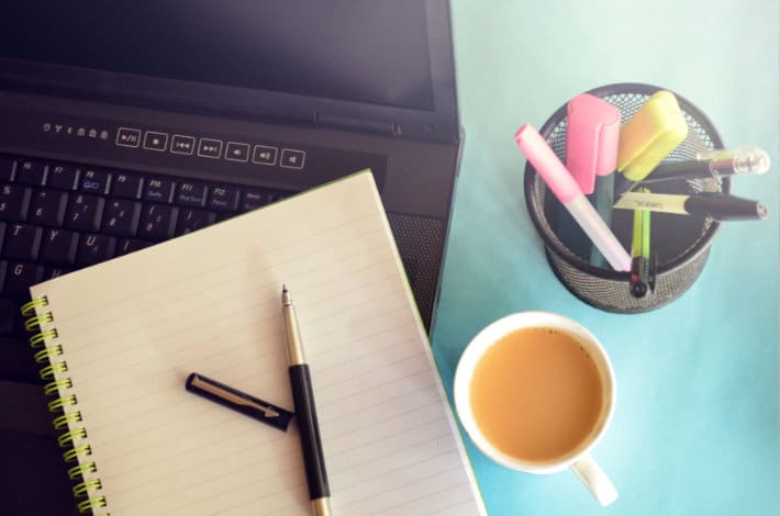 a laptop, notebook, pen, pen holder full of writing utensils, and a coffee cup sit on a blue desk