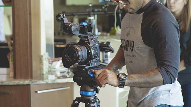 a man with forearm tattoos and rolled up sleeves operates a camera on a tripod