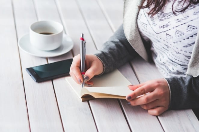 a woman writes in a notebook on a white desk next to her phone and a half-full coffee mug