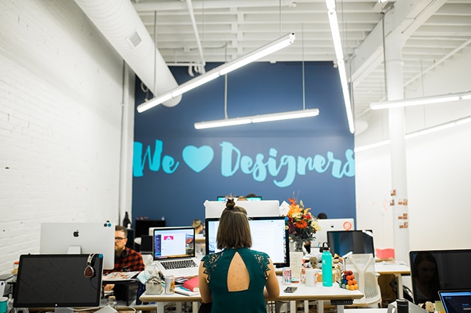 layout by flywheel design perfect website navigation woman at desk in front of big blue wall with we heart designers painted on it
