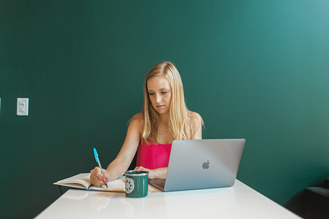 woman writing content marketing ideas in notebook with laptop and flywheel mug nearby